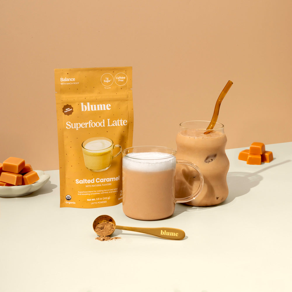 A packet of Blume Superfood Latte mix is displayed alongside a clear mug of frothy latte and a wavy glass with iced latte, straw included. A spoon with powder and a bowl of caramel cubes are also shown against a beige background.